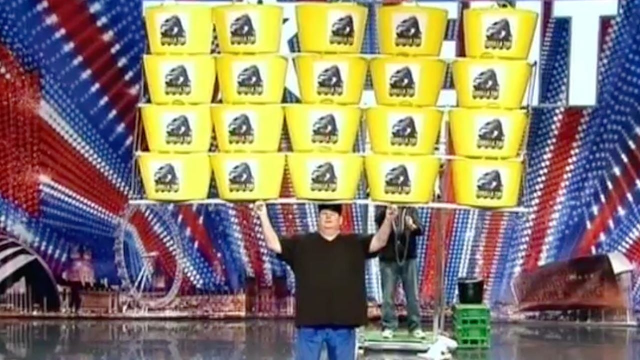 A person stands on stage holding a large rack stacked with yellow buckets in a pyramid shape, with a colorful background behind them.