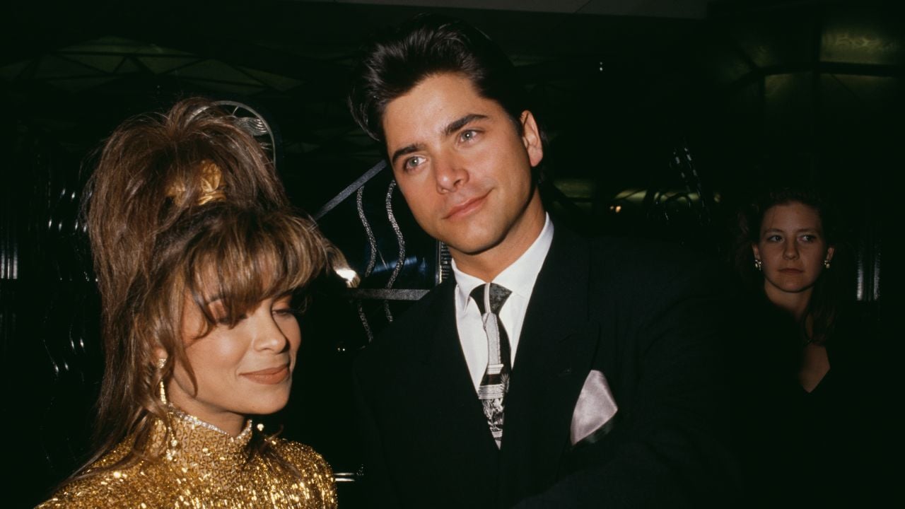 A woman in a gold sequin dress stands beside a man in a black suit and patterned tie at a formal event, with another woman in the background.