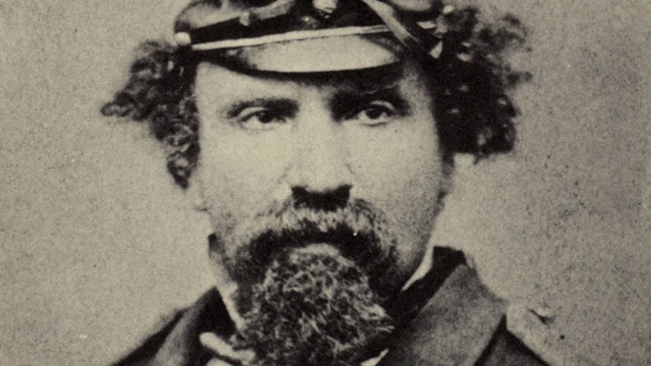 A black-and-white portrait of a man with curly hair, a full beard, and mustache, wearing a uniform cap and dark clothing, looking directly at the camera.