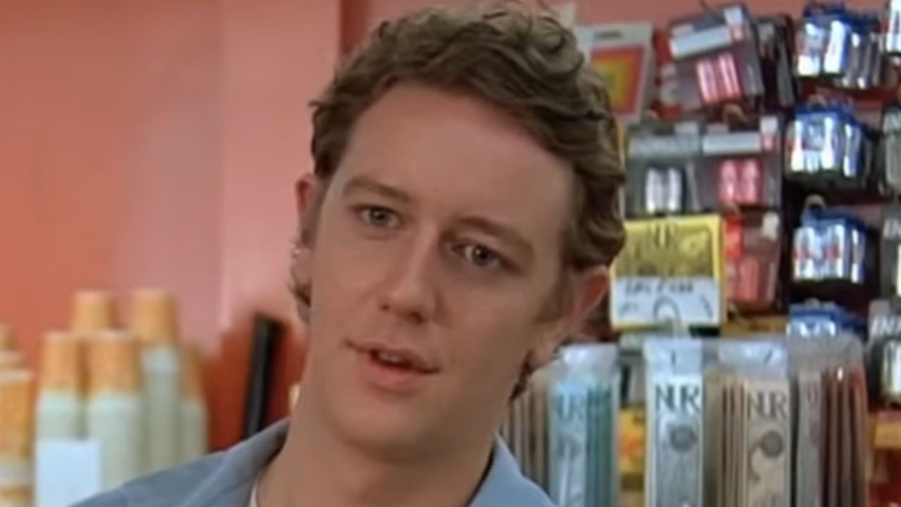 A young man with curly brown hair stands in a store aisle with various products displayed behind him, including drink cups and hanging items on racks.