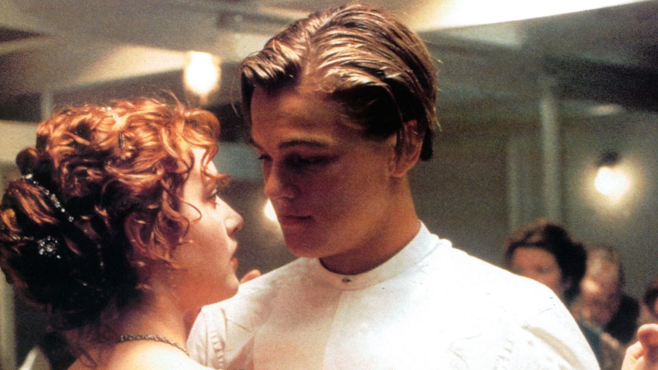 A young couple dressed in period clothing share an intimate moment, gazing into each other’s eyes while slow dancing at a formal event.