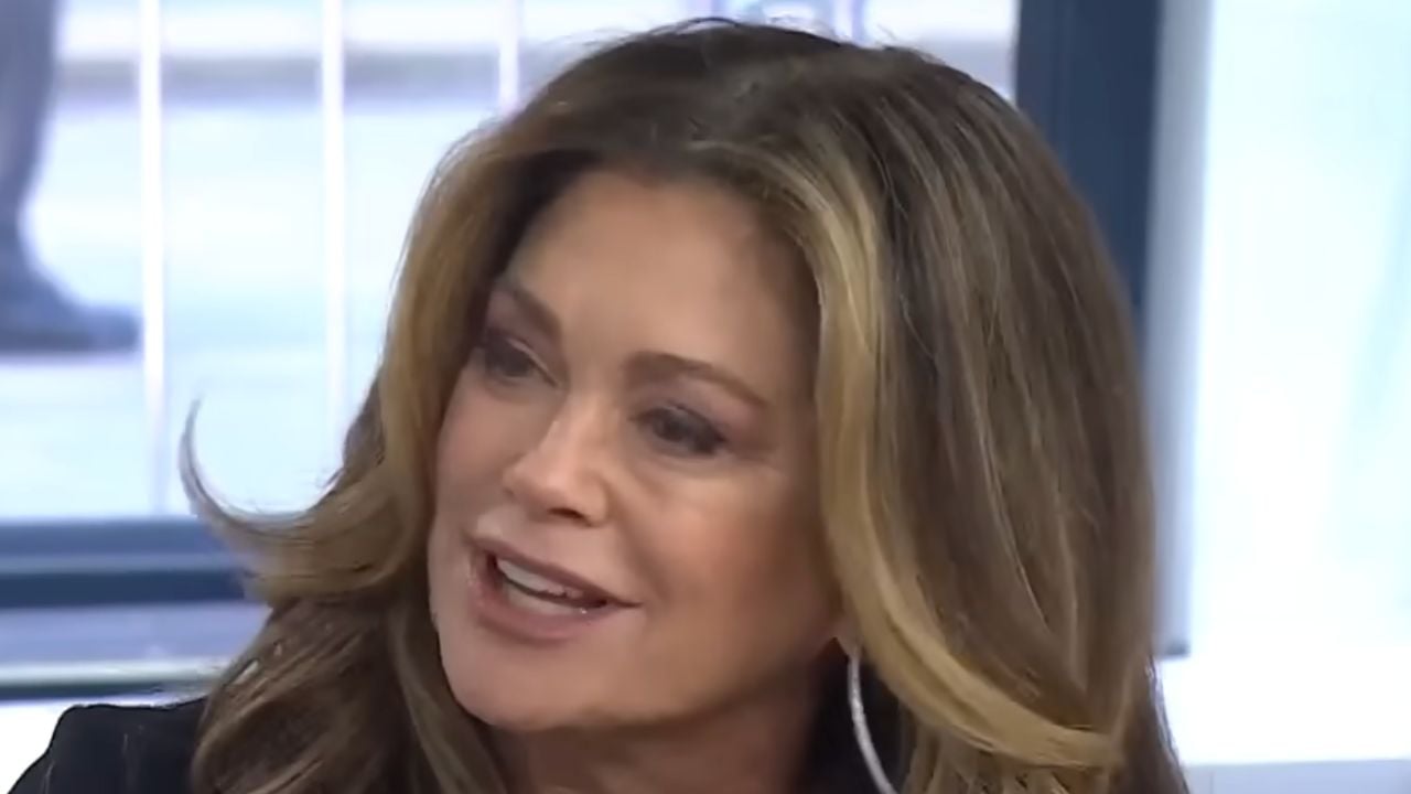 A woman with long, wavy brown hair and large hoop earrings is smiling and talking indoors, with a blurred window and light background behind her.