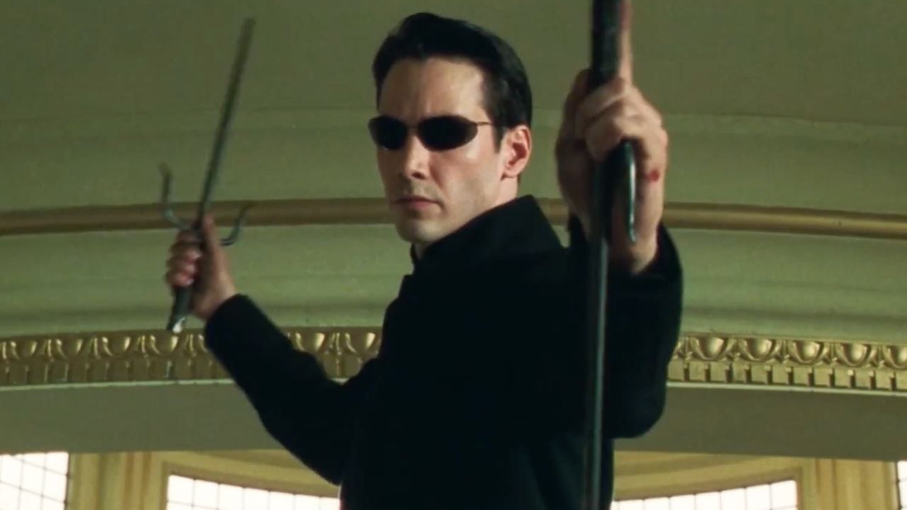 A man in black sunglasses holds two weapons, poised for action, in an ornate room with cream-colored walls and gold trim.
