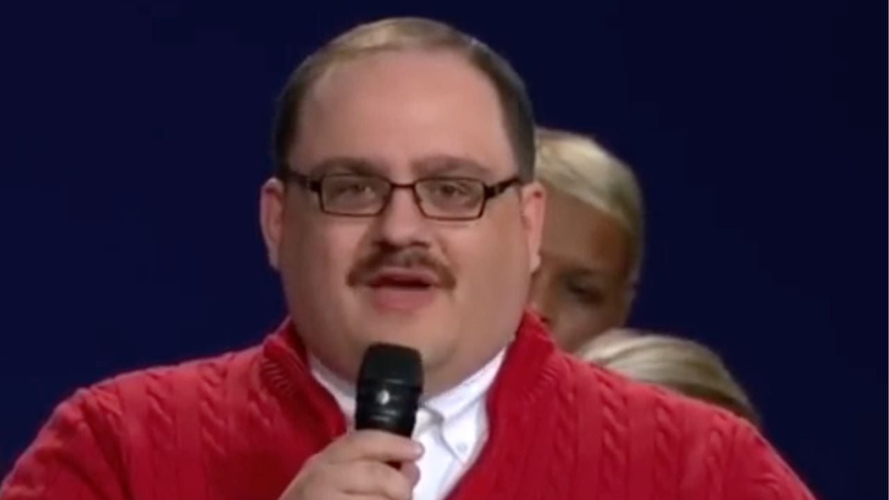 A man with glasses, a mustache, and short hair wears a red sweater and holds a microphone, speaking. There are blurred people in the background.