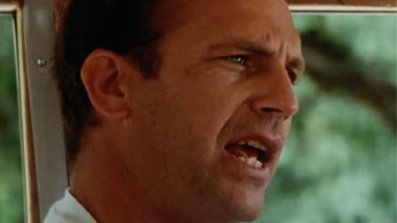 A man with short brown hair is speaking, shown in close-up, with a blurred green background behind him. He appears to be outdoors and is wearing a light-colored shirt.