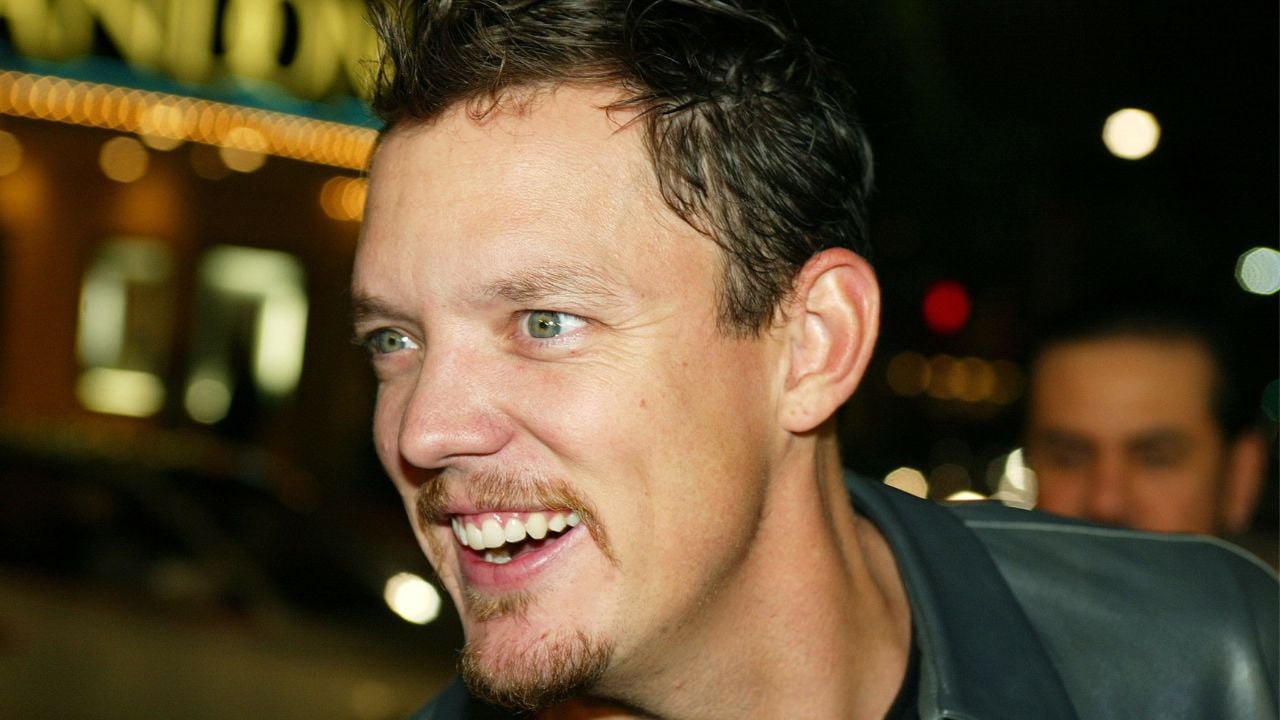 A man with short dark hair, light eyes, and a light goatee smiles at night. Bright lights and blurred signs glow in the background.