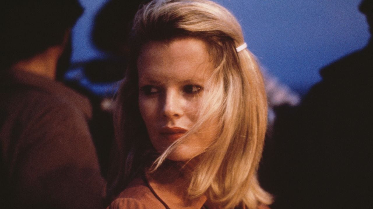 A woman with blond hair, wearing a red top, looks over her shoulder with a serious expression. The background is dark and blurry, suggesting an outdoor or evening setting.