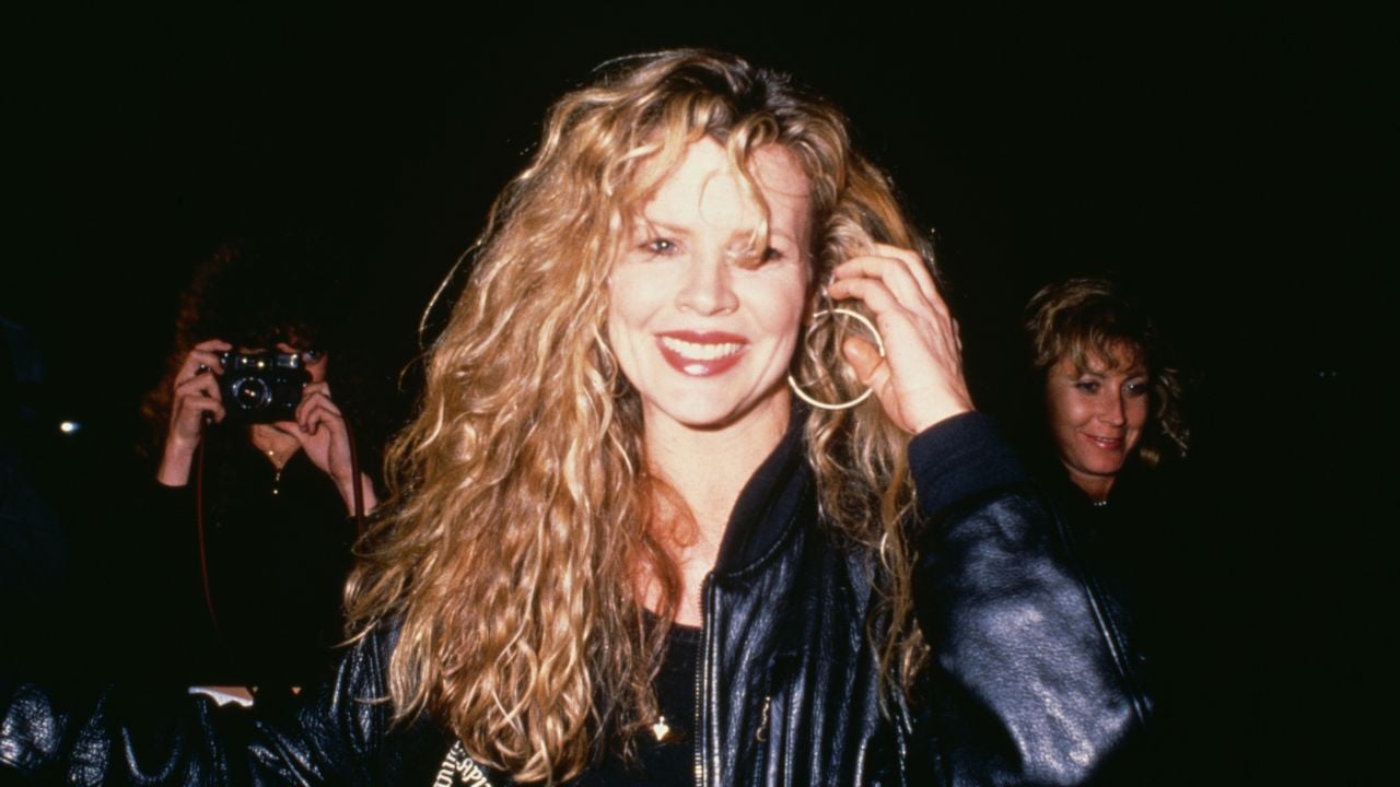 A woman with long, wavy blonde hair smiles at the camera, wearing a dark jacket and large hoop earrings. People in the background, one holding a camera, are slightly out of focus.