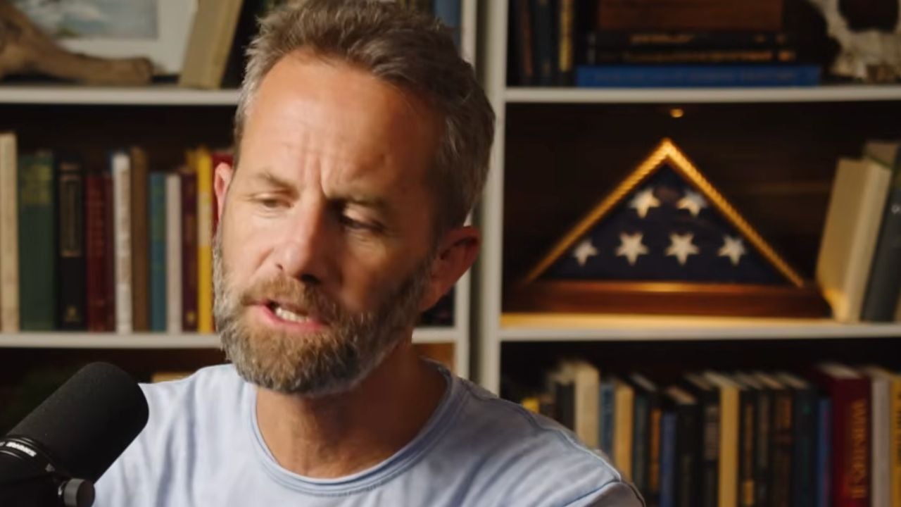A man speaking into a microphone in front of bookshelves, with a folded American flag displayed behind him.