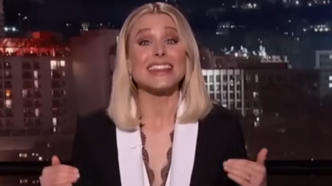 A woman with blonde hair in a black and white outfit smiles and gestures with both hands while speaking on a nighttime TV show set. City lights and buildings are visible in the background.