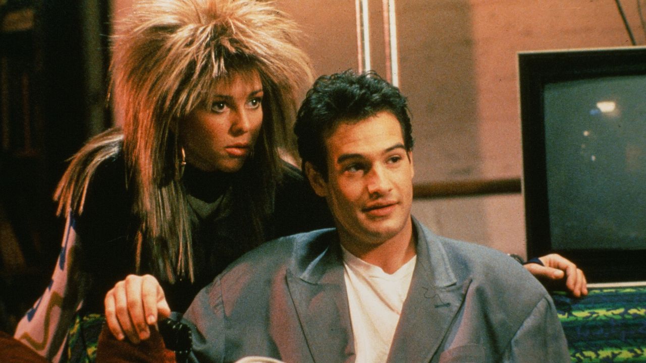 A woman with spiky hair and bold makeup leans toward a man in a loose jacket sitting on a couch. There is a TV and colorful patterned blanket in the background.