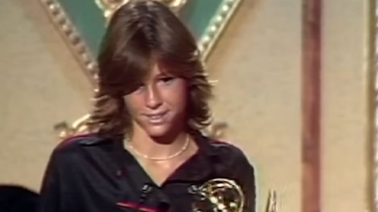 A young person with shaggy brown hair and a black shirt stands holding a golden trophy, with an ornate gold and green background behind them.