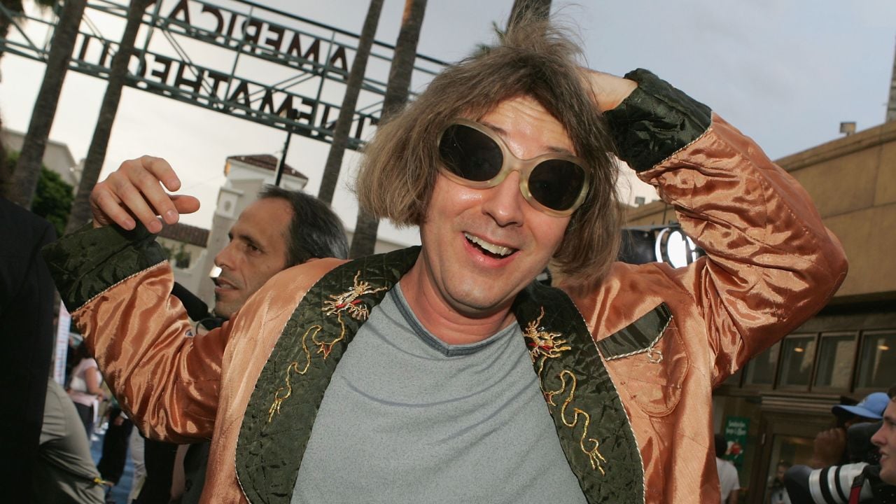 A person wearing round sunglasses and a shiny patterned jacket smiles and poses with one hand on their head at an outdoor event, with palm trees and a sign in the background.