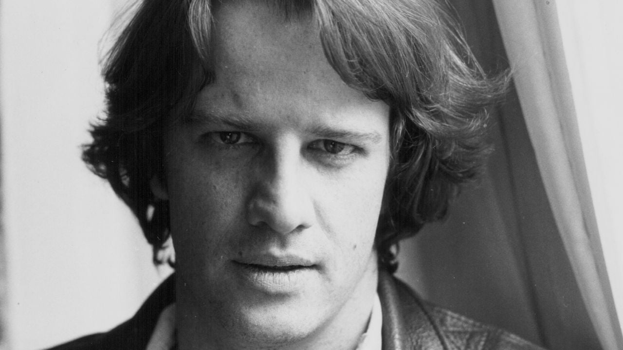 A black and white close-up portrait of a person with medium-length hair, looking into the camera with a neutral expression, wearing a leather jacket and a light shirt, standing near a curtain.
