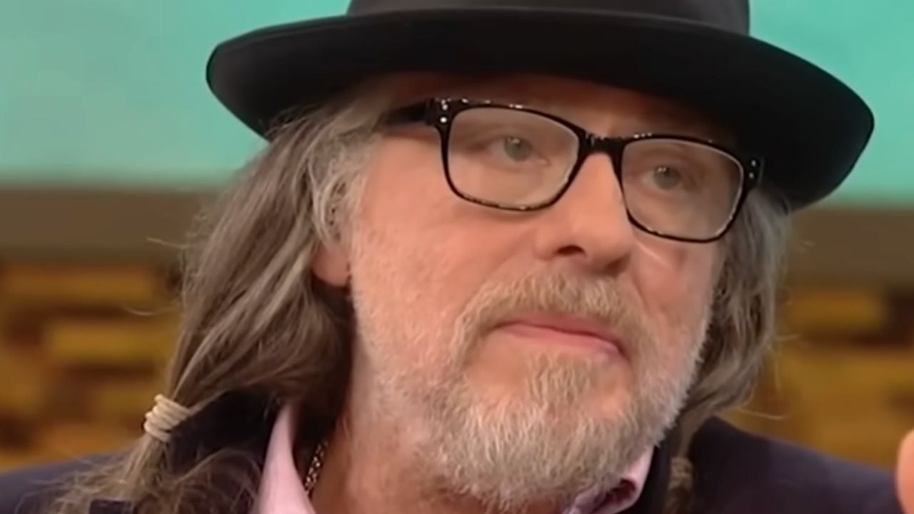 A person with long gray hair, a beard, glasses, and a black hat is looking slightly to the side. The background is blurred, with warm and teal tones.