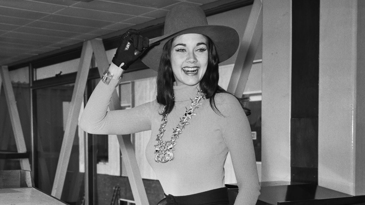 A smiling woman in a wide-brim hat, long-sleeve top, gloves, and large necklace poses indoors, lifting her hat with one hand.