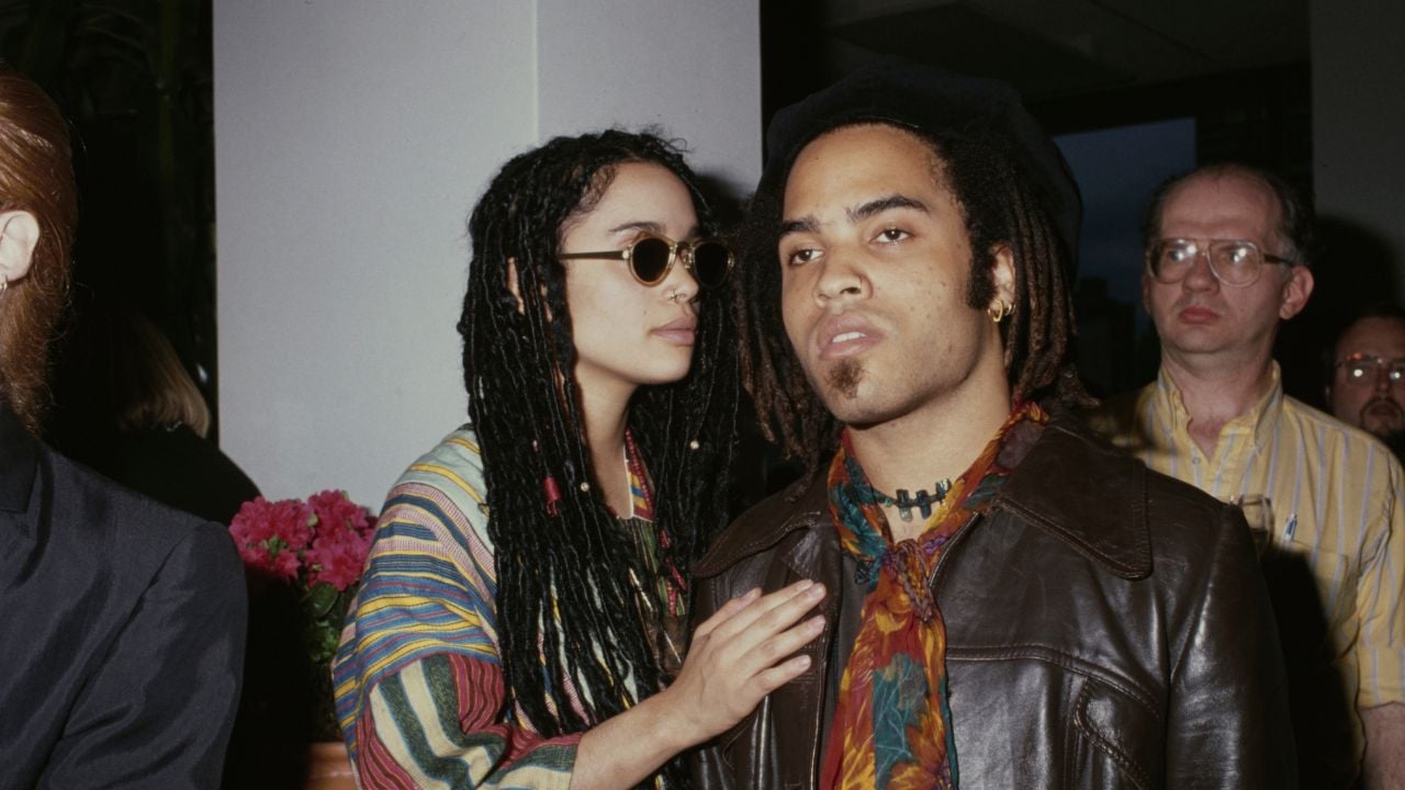 Two people with dreadlocks, one wearing sunglasses and colorful clothing, stand close together indoors. The other wears a leather jacket and hat. Other people and flowers are visible in the background.