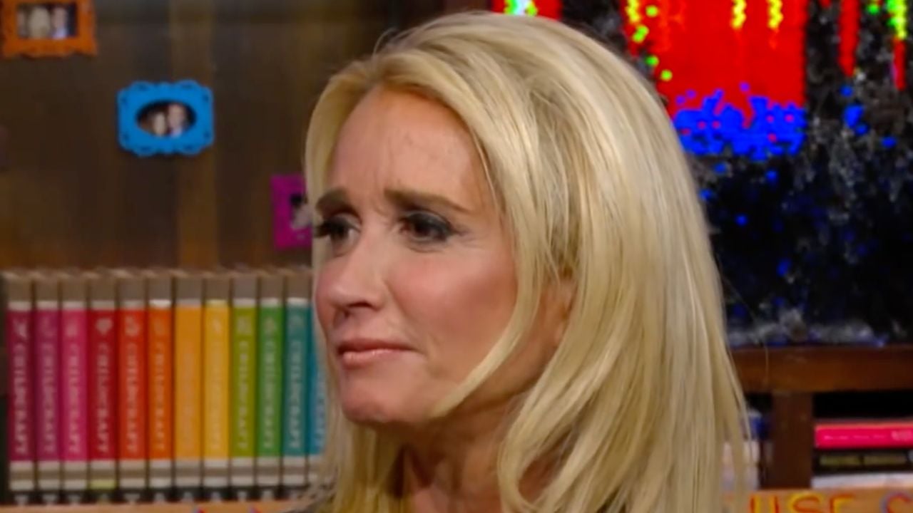 A woman with long blonde hair looks to the side in a colorful room with books and decorative frames in the background.