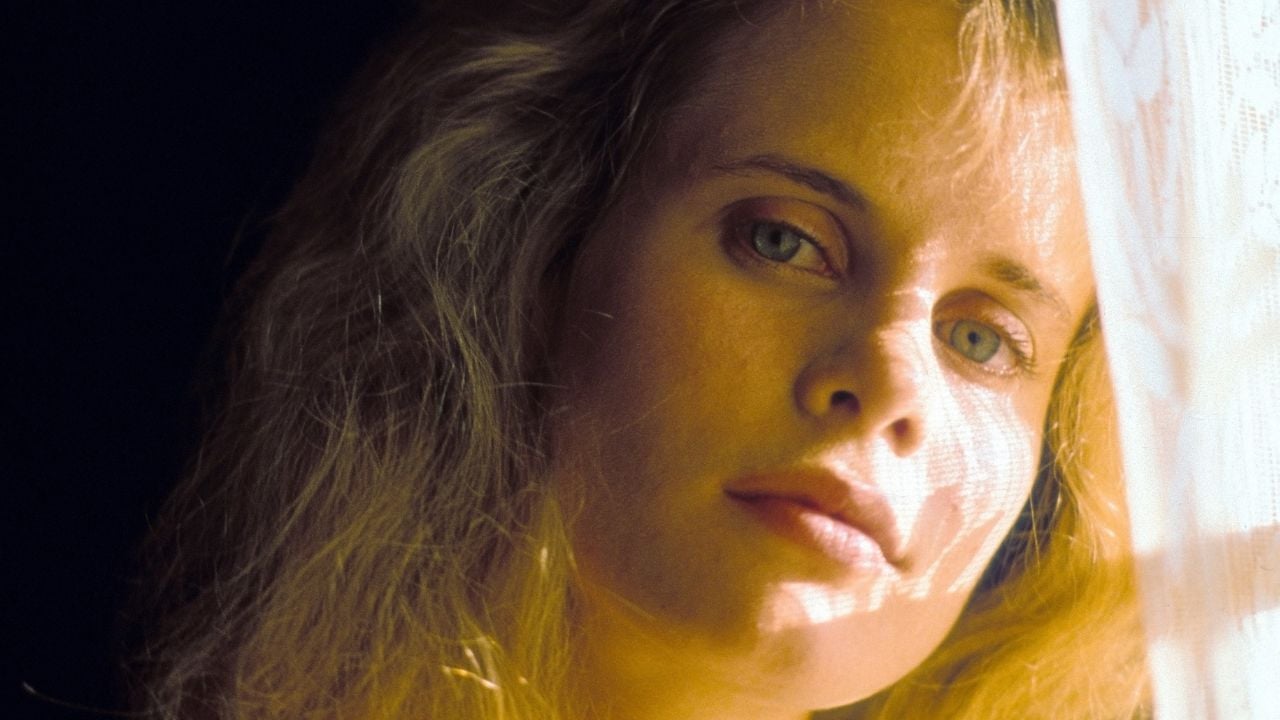 A woman with blonde hair gazes softly toward the camera, sunlight filtering through a lace curtain partially illuminating her face, creating gentle shadows.