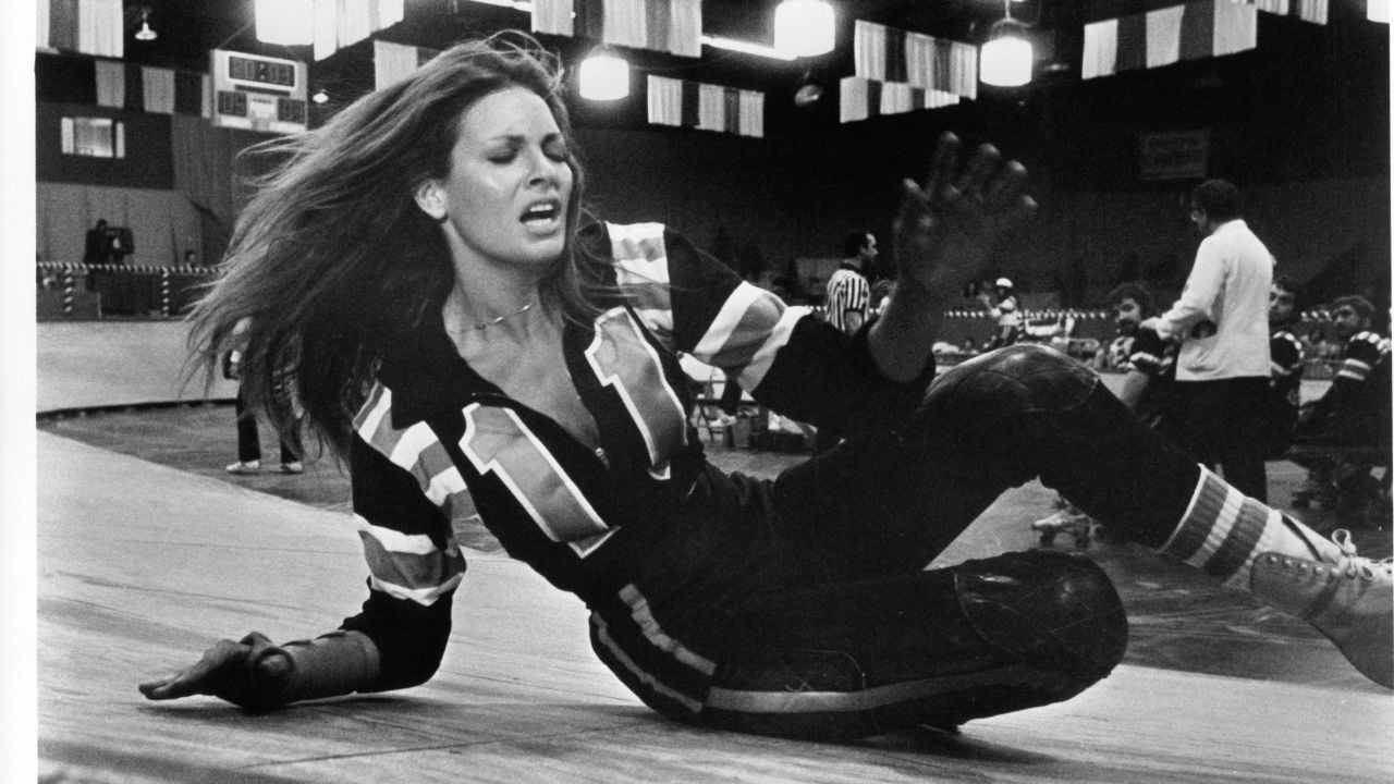 A woman in a roller derby uniform with the number 11 falls on a rink, looking distressed. Other players and referees are visible in the background.