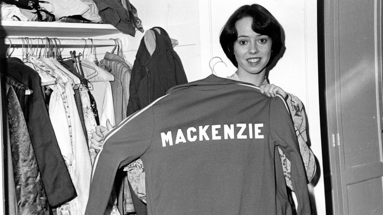 A woman smiles and holds up a jacket with “MACKENZIE” written on the back. She stands beside an open closet with hanging clothes. The image is in black and white.