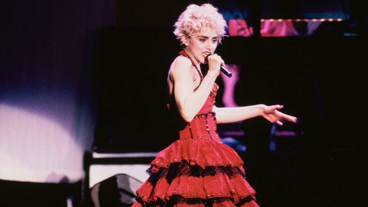 A singer with short, curly blond hair performs on stage wearing a layered red dress, holding a microphone and gesturing with her hand, with a dark background behind her.