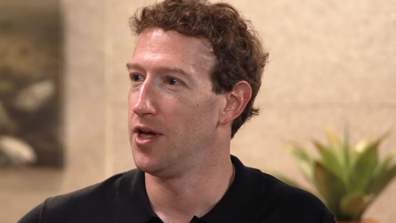 A man with short curly brown hair wearing a black polo shirt speaks while looking to the side. There is a plant and a blurred painting in the background.