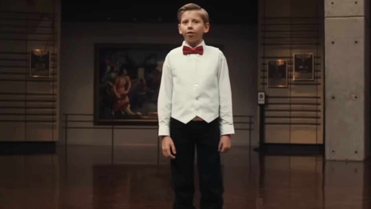 A young boy in a white vest, red bow tie, and black pants stands in an art gallery with paintings on the walls behind him.