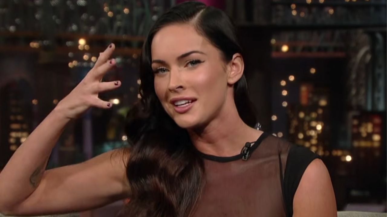 A woman with long dark hair, wearing a sheer black top, gestures with her hand next to her face while speaking on a talk show set with blurred lights in the background.