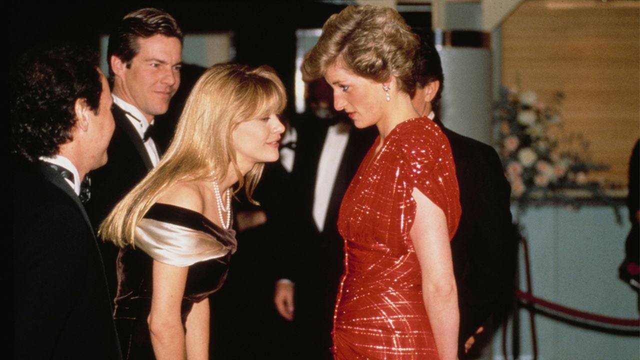 A woman in a red dress faces a woman in an off-shoulder gown, both making eye contact at a formal event. Two men in tuxedos stand nearby. The background includes flowers and elegant decor.
