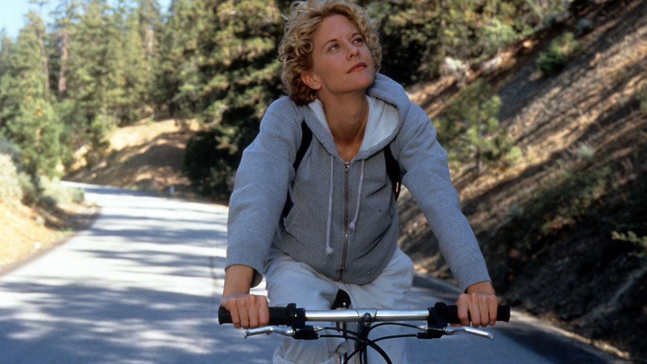 A person with short curly blond hair rides a bicycle on a sunny road through a forest, wearing a light gray hoodie and white pants, looking up thoughtfully.