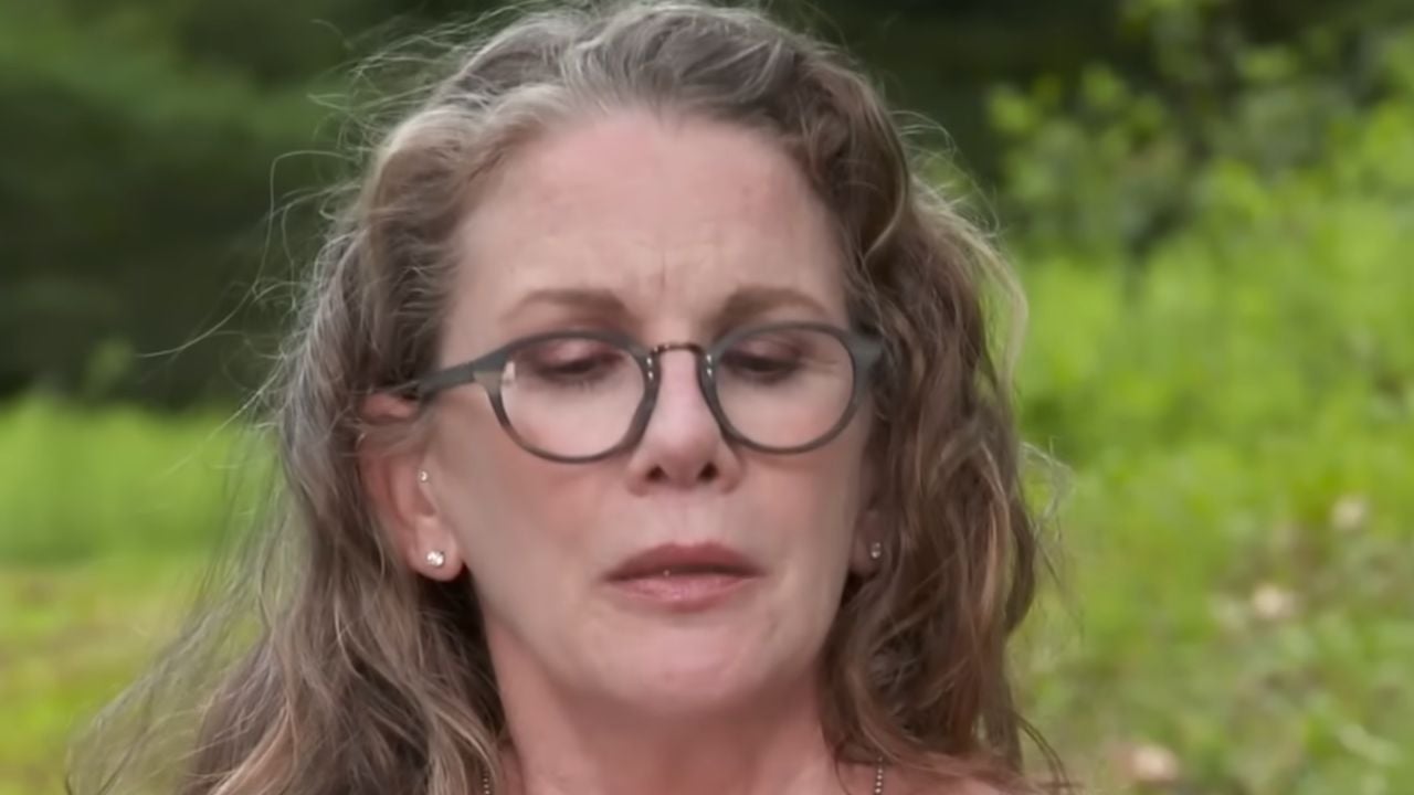 A woman with long wavy hair and glasses is outdoors, looking down with a serious expression. Green foliage and blurred trees are visible in the background.