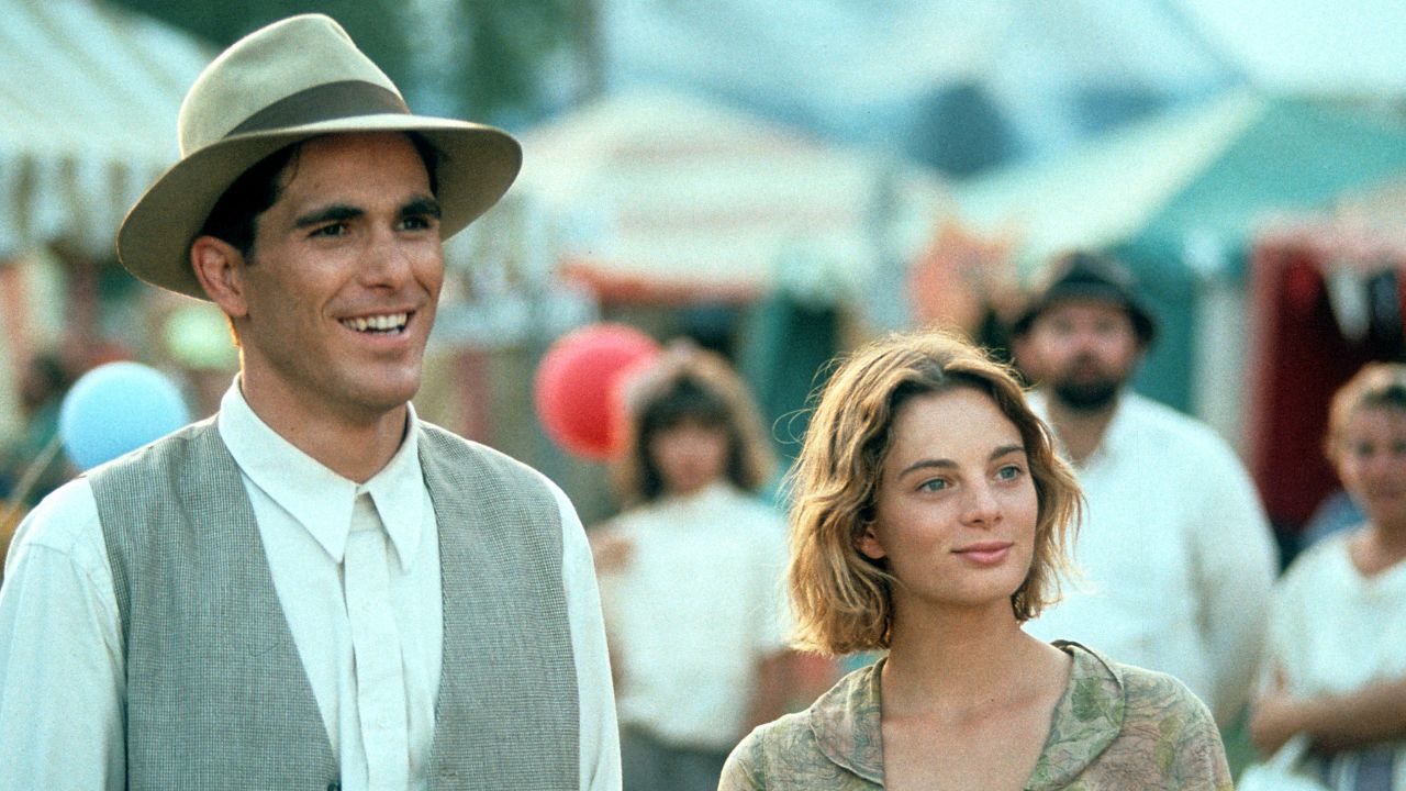 A man in a hat and vest stands next to a woman with short hair at an outdoor fair, smiling. People and colorful tents are visible in the blurred background.