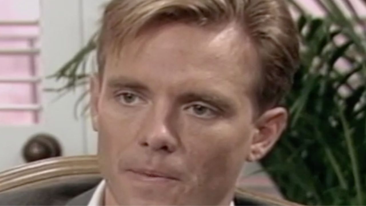 A person with short light brown hair and fair skin looks serious, sitting indoors with a blurred plant and window in the background.