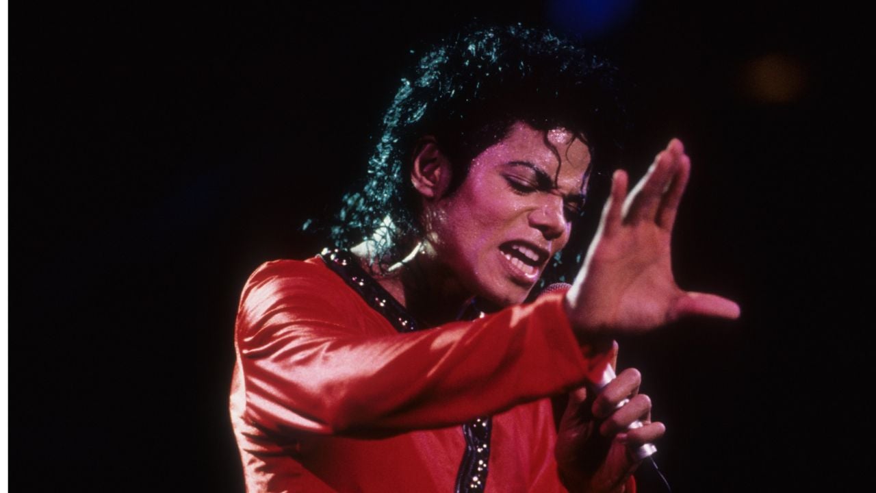 A singer with curly black hair in a red outfit passionately performs on stage, holding a microphone and raising one hand in front of their face against a dark background.