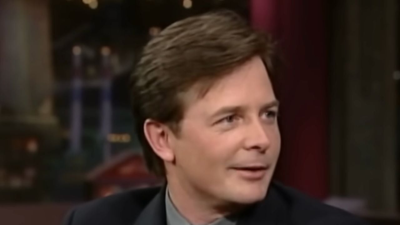 A man with light skin and brown hair is wearing a dark suit and looking to his left. The background is blurred with warm lighting.