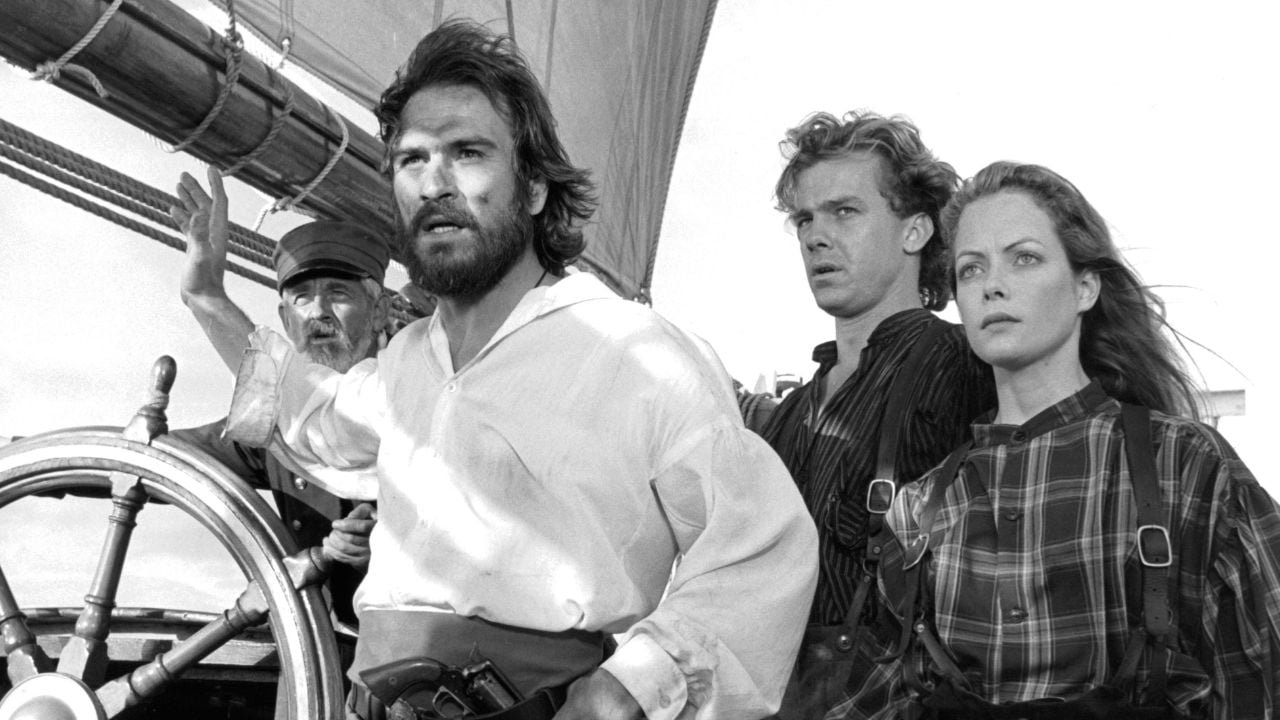 Three people in old-fashioned clothing stand on a ship’s deck, looking intensely ahead. A man at the ship’s wheel is behind them. The scene appears dramatic and serious, suggesting an adventure or historical setting.