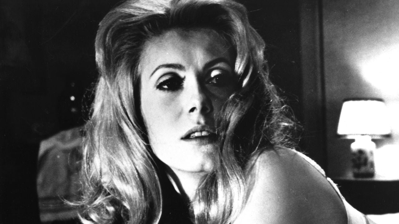 Black and white photo of a woman with voluminous, wavy hair looking slightly to the side. She has dramatic eye makeup and a soft expression. A lamp and part of a wall are visible in the blurred background.
