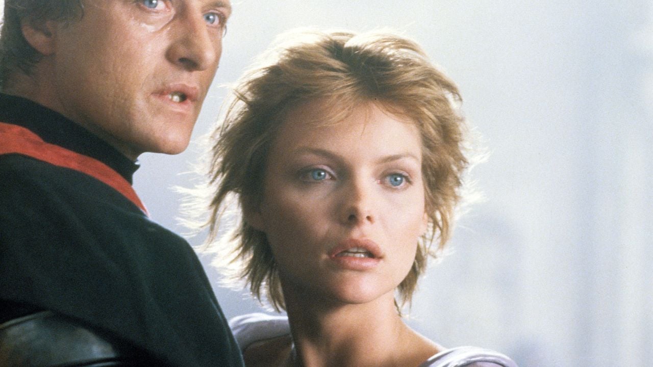 A man in a dark outfit stands close behind a woman with short blonde hair. Both look off-camera with serious, intense expressions against a softly lit background.