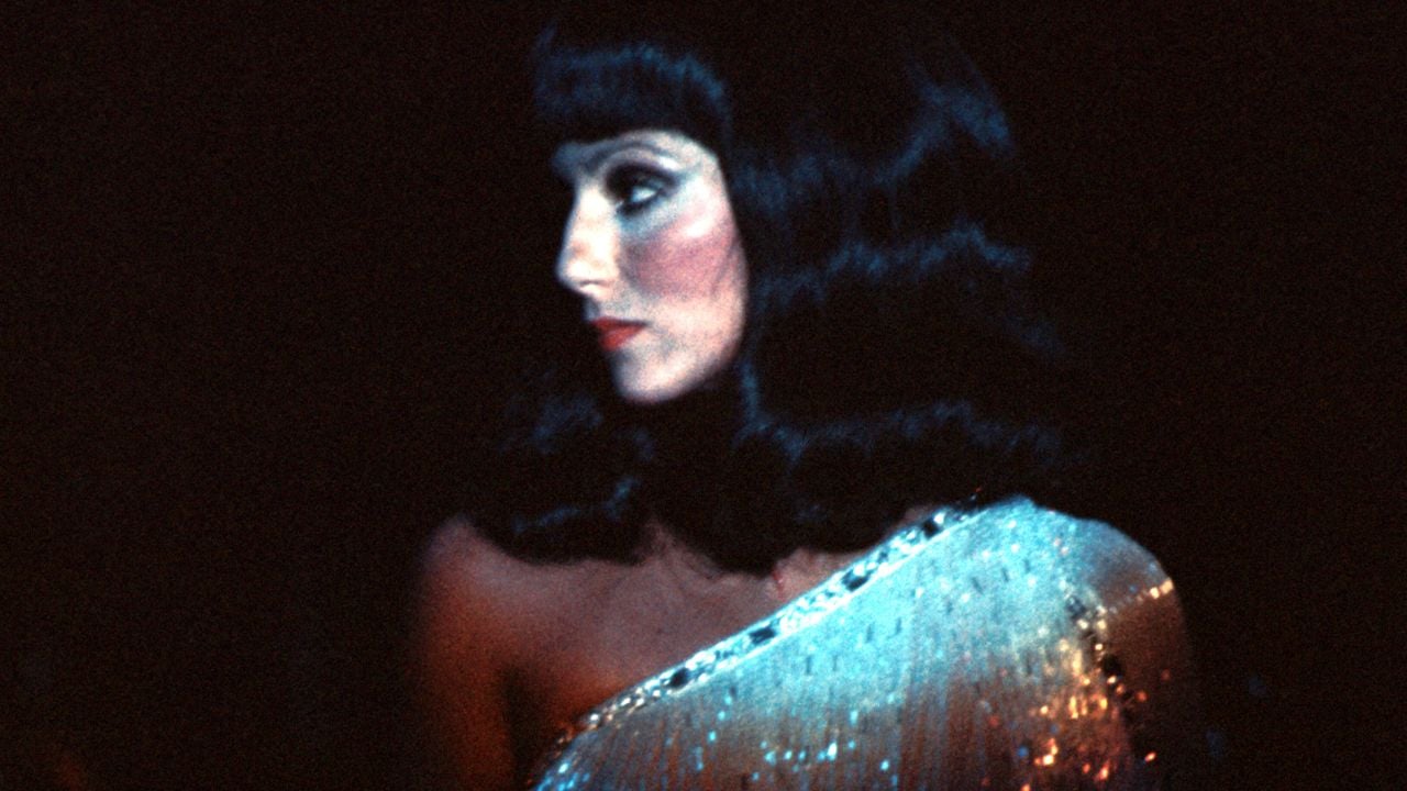 A woman with long dark hair and bangs in profile wears a sparkling, one-shoulder dress against a dark background.