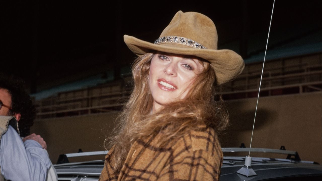 A woman with long, wavy hair wears a brown plaid coat and a tan cowboy hat with a decorative band, smiling outdoors at night.