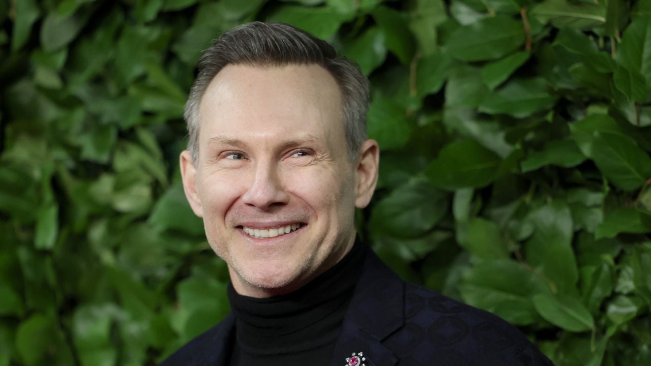 A smiling man with short, light brown hair wearing a black turtleneck and blazer stands in front of a lush green leafy background.