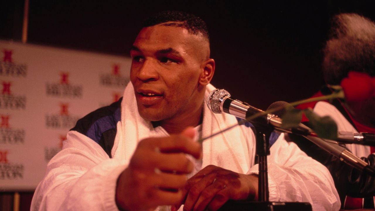 A man in a white jacket sits at a press conference table, holding a red rose near a microphone, with a towel draped around his neck and a branded backdrop behind him.