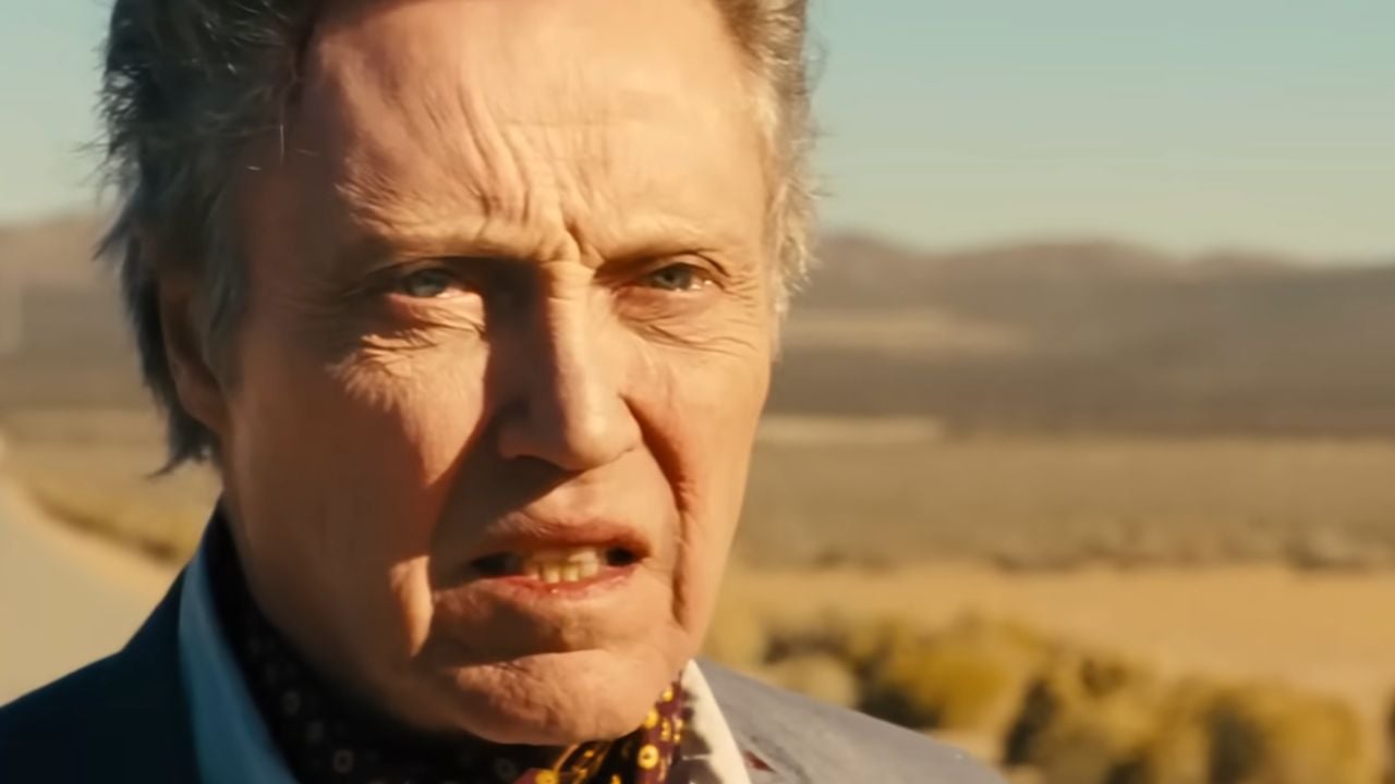 An older man with light hair and deep wrinkles looks concerned, standing outdoors with a blurred desert landscape and mountains in the background.