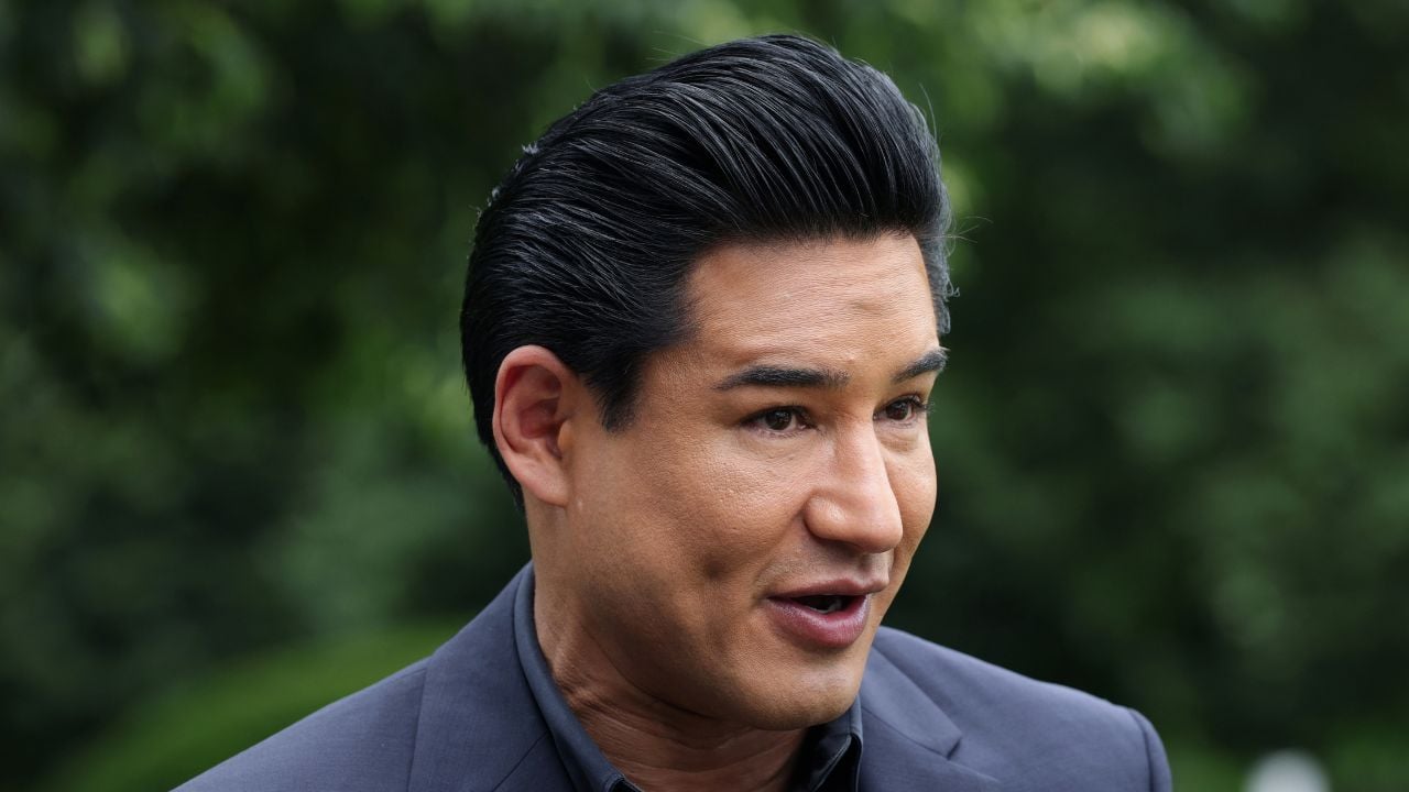 A man with slicked-back black hair wearing a dark suit speaks outdoors with greenery blurred in the background.