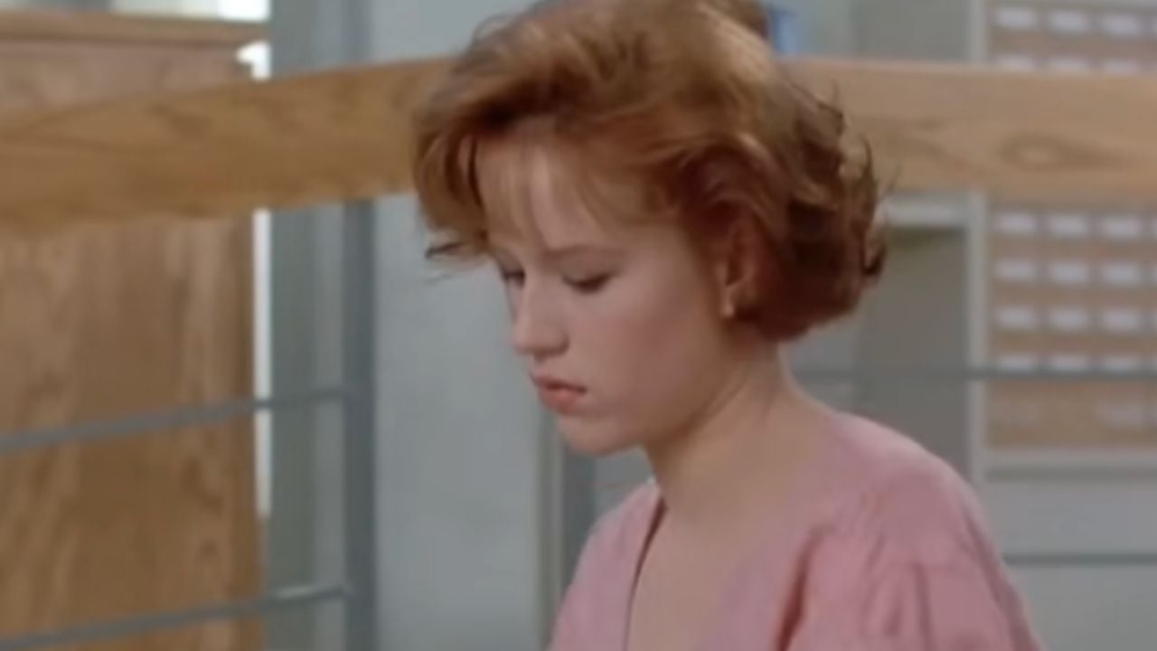 A young woman with short, wavy red hair wearing a pink top looks down with a serious expression in a modern indoor setting with light wood and metal details.
