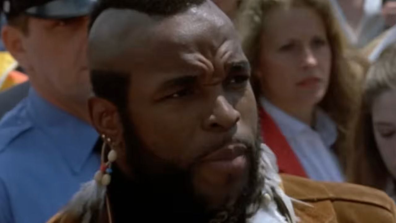 A man with a mohawk hairstyle, beard, and feather earring stands outdoors among a crowd, including a uniformed police officer and several people in the background.