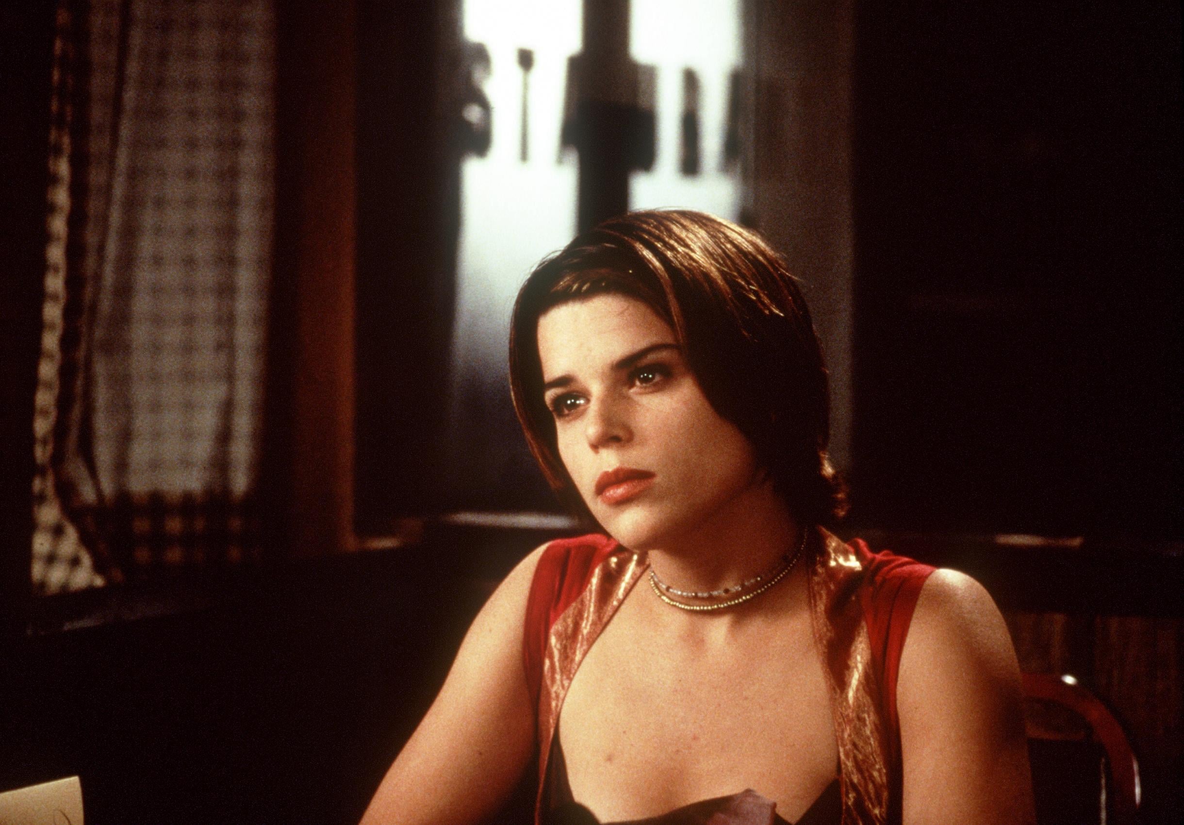A woman with short brown hair and a red top sits at a table, looking thoughtful. The background is dimly lit, with a blurred window and part of a sign visible behind her.