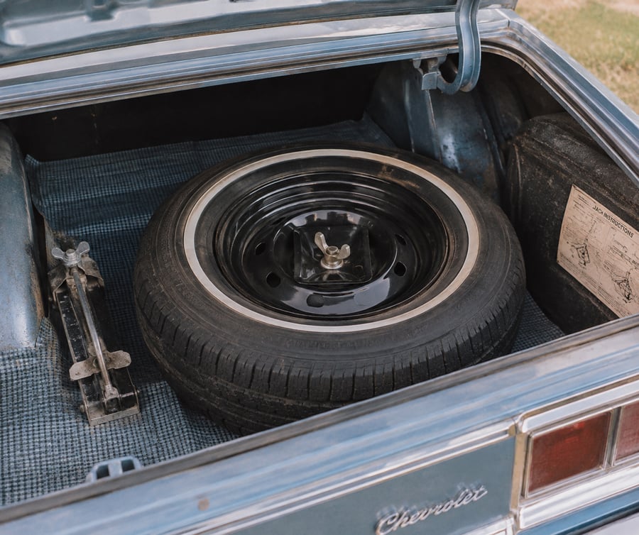 Open trunk of a classic Chevrolet car showing a spare tire, a metal car jack, and an instruction label attached to the inside of the trunk lid. The trunk lining has a blue checkered pattern.
