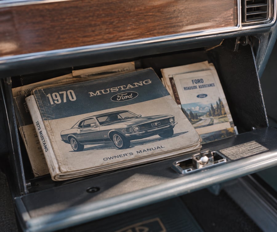An open glove box showing a 1970 Ford Mustang owner’s manual and other booklets inside a classic car with a wood-trimmed dashboard.
